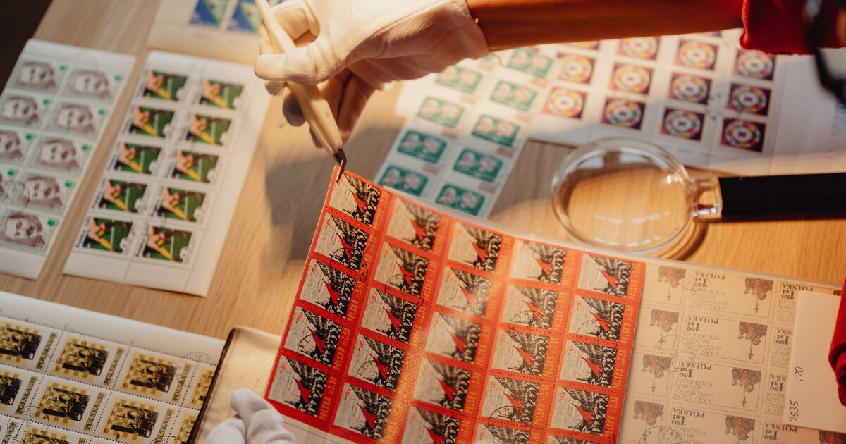 A collector in white gloves examining stamp sheets with tweezers and a magnifier